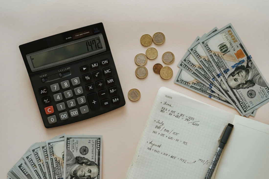 Black calculator, dollar bills, open notebook, and pen on desk