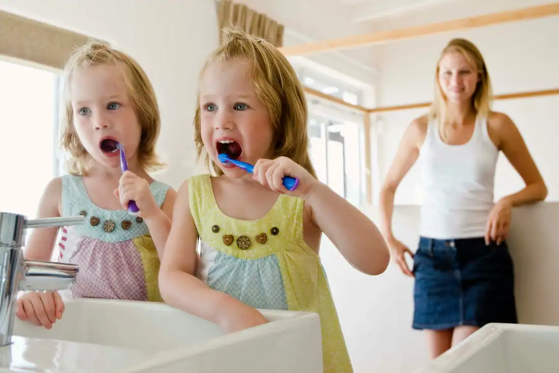 two girls brushing their teeth with their mom watching